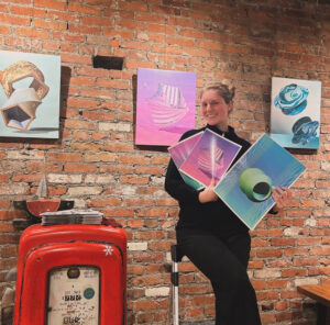 Artist Anna Geiger holding several prints of their art in front of a brick wall at Diesel Cafe.